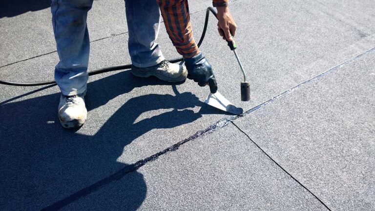 Homeowner comparing roof color samples beside siding and trim on a residential house.