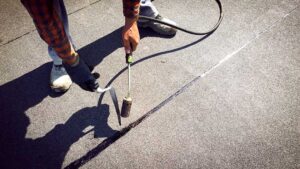 Person clearing leaves from a gutter beside a shingled roof with a ladder and downspout visible.