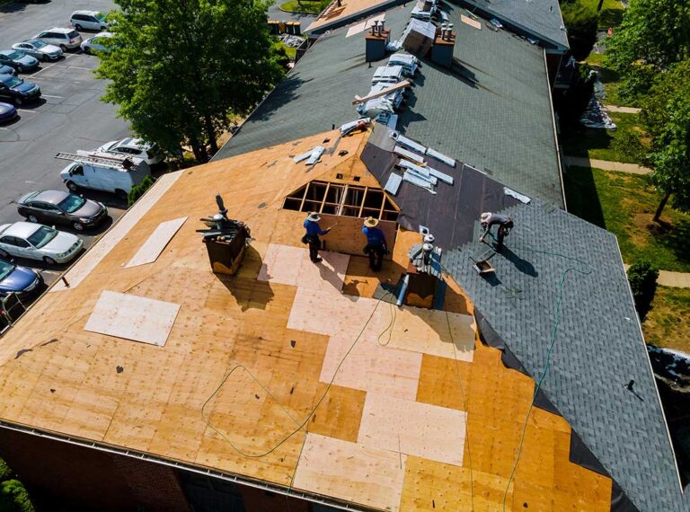 Person inspecting roof shingles and gutters beside a two-story home in autumn.