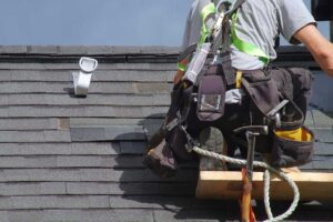 Person clearing leaves from a residential gutter beside a shingled roof with a ladder on the ground.