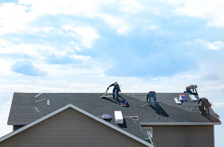A homeowner inspects a snow-covered roof and cleared gutters from the ground beside a winter house.
