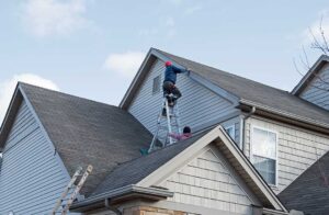 Roofing workers replace shingles above a home while dry attic insulation remains visible below.