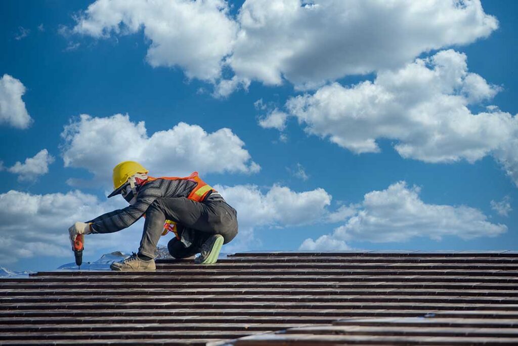 Homeowner viewing roof shingles and clearing leaves from a gutter beside a two-story house.