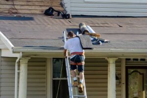 A homeowner reviews roof plans beside a snow-covered house while a code inspector examines the roof structure.