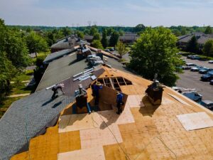 Workers remove old shingles beside a house while exposed roof decking is inspected and debris is contained below.